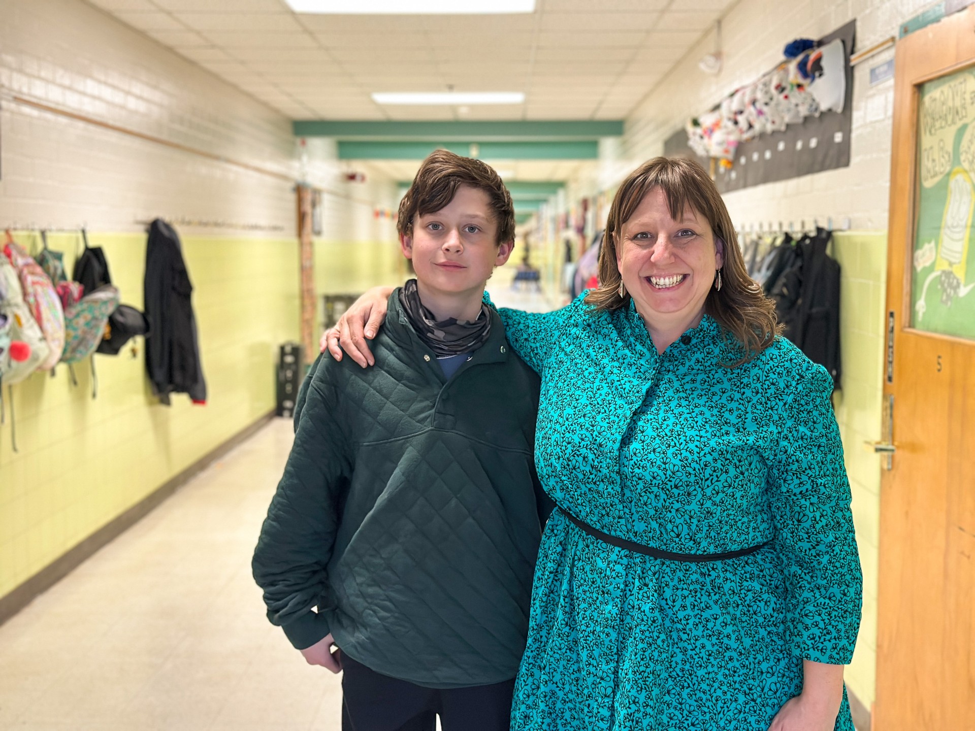 A woman in a blue dress stands with her arm around her young son in the school hallway