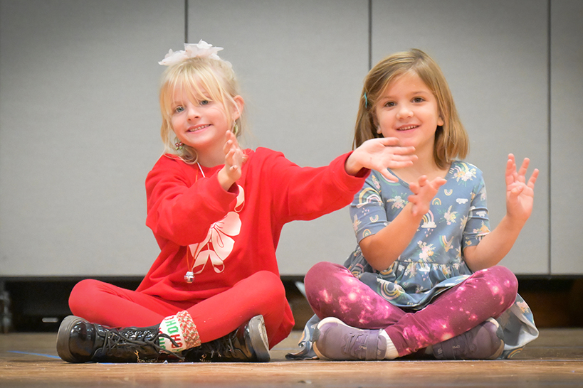 Two little girls sit on the stage waving their arms and smiling