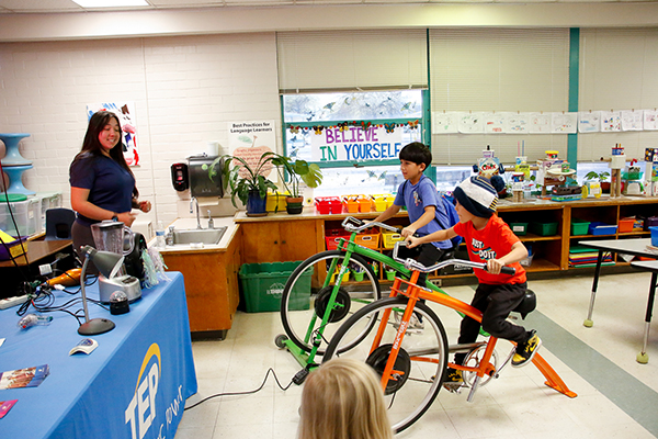 A woman smiles as two boys pedal bicycles in a classroom