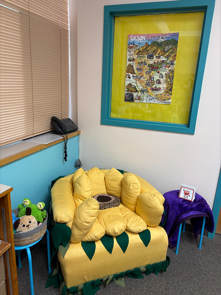 A yellow sunflower-shaped chair in a corner with a basket of stuffed animals, a table with a purple blanket on it and a phone on a shelf
