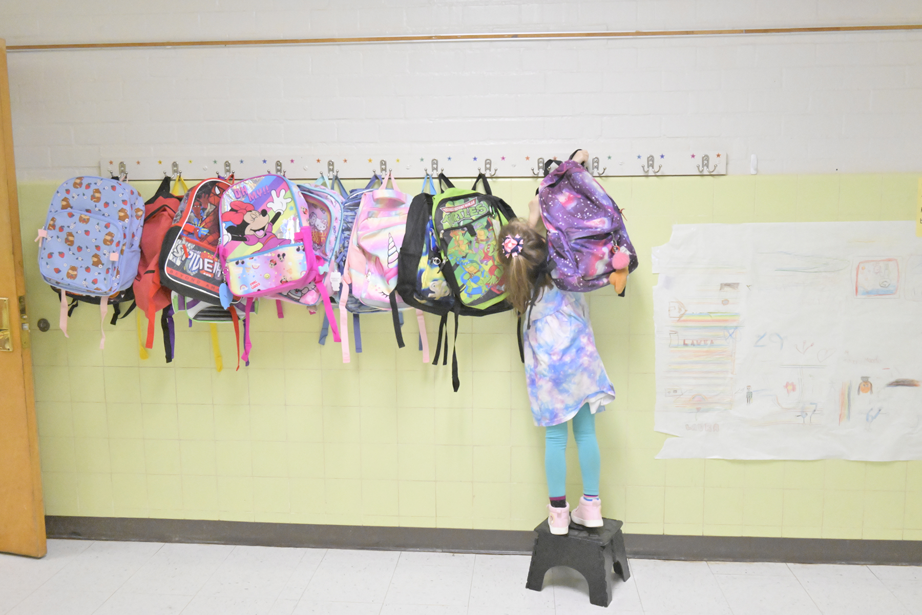 A little girl stands on a stool to reach her backpack